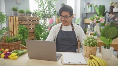 Canlı bir çiçekçide dizüstü bilgisayar kullanan gülümseyen Asyalı adam bitkiler ve çiçeklerle çevrili..