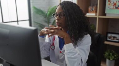 Kıvırcık saçlı, yorgun ve genç bir Afro-Amerikan kadın doktor kapalı bir klinikte bilgisayarının başında çalışırken gözlüğünü çıkartıyor..
