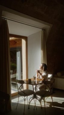 Güneşli bir kafeteryada oturan kadın, kahve içiyor, açık hava bahçesiyle geniş bir pencereden görülüyor, rahat bir atmosferi ve rahat bir ortamı ön plana çıkarıyor..