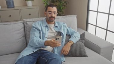 Genç adam modern bir oturma odasındaki akıllı telefonu kullanarak kanepede dinleniyor. Arka planda bitkiler ve dekorlar var. Gözlük takıyor ve rahat görünüyor.