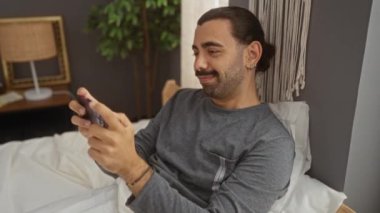Bıyıklı genç adam rahat bir yatak odasında arka planda bitkilerle otururken akıllı telefon kullanıyor.