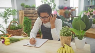 Genç Asyalı erkek çiçekçi bir telefon görüşmesi yapıyor ve parlak bir dükkanda taze çiçeklerle çevrili emirler yazıyor..