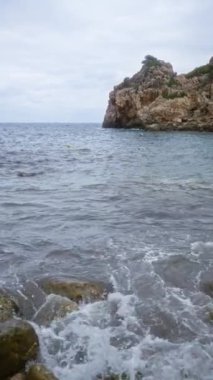 Mallorca 'daki Cala Deia' da kayalara çarpan dalgalar ve bulutlu bir gökyüzünün altındaki uzak deniz manzaralı bir sahil şeridi.