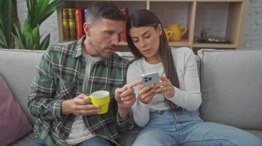 İspanyol bir çift evde uzanıyor, kahve içen bir adam ve akıllı telefon kullanan bir kadın, gri bir kanepede rahatça oturuyorlar..