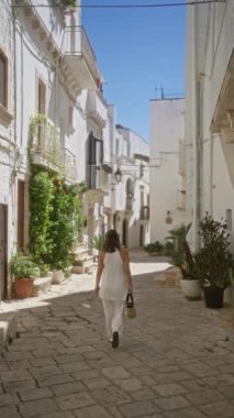 Genç bir kadın güneşli bir günde beyaz binalar ve yemyeşil bitkilerle çevrili Locorotondo, Puglia, İtalya 'nın büyüleyici eski sokaklarında gezinir..  