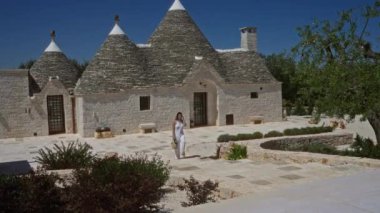 Beyaz elbiseli genç İspanyol kadın açık mavi Avrupa gökyüzü altında eşsiz bir mimari sergileyen Alberobello, Puglia, İtalya 'daki geleneksel taş trulli evlerin yanında duruyor..