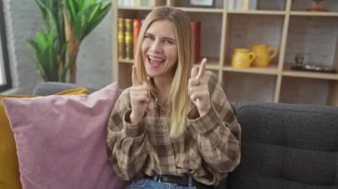 Evdeki kanepede oturan neşeli, genç sarışın kadın. Parıldayan bir gülümsemeyle kamerayı işaret ediyor. Sevincin, iyi hislerin ve mutluluğun büyüleyici portresi..