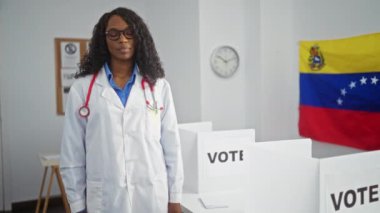 Kıvırcık saçlı ve gözlüklü Afrikalı genç bir Amerikalı kadın, arka planda Venezuela bayrakları ve oy kullanma kabinleriyle kapalı bir seçim ortamında kameraya gülümsüyor..