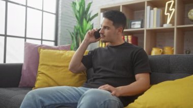 Modern bir oturma odasında gri bir koltukta oturan düşünceli bir adam telefonda konuşuyor.