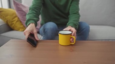 Sarı kupa ve akıllı telefon tutan kel bir adam, kapalı bir gri koltukta oturuyor renkli yastıklarla çevrili..