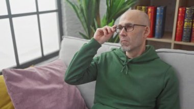 Kapalı bir koltukta, renkli yastıklar ve kitaplıkla çevrili bir şekilde otururken sakallı, dalgın, kel bir adam gözlüğünü çıkarıyor..
