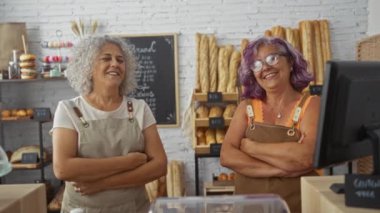 İki olgun kadın fırıncı kollarını kavuşturup gülümseyerek taze ekmek ve hamur işleriyle dolu kapalı bir fırında duruyorlar.