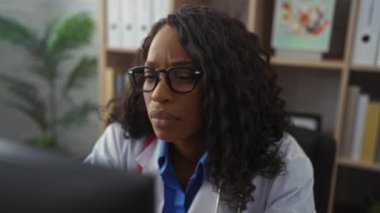 Kıvırcık saçlı ve gözlüklü güzel bir Afro-Amerikalı kadın, kapalı bir klinikte steteskop ve kitaplık arka planıyla çalışıyor..