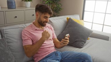 Oturma odasında telefona gülümseyen genç adam koltukta heyecan sergiliyor.