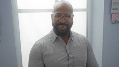 Kapalı ofis ortamında gülümseyen sakallı ve gözlüklü genç Afrikalı Amerikalı bir adam profesyonel ve misafirperver bir tavır sergiliyor..