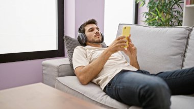 Sakallı genç bir İspanyol, akıllı telefon kullanırken ve kulaklık takarken koltukta dinleniyor..