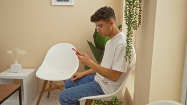 Young hispanic man sitting in a modern waiting room, focused on his phone, wearing casual clothes and glasses, surrounded by white furniture and indoor plants.