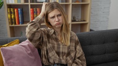 Kafası karışmış genç bir kadın modern bir oturma odasında düşünceli bir ifadeyle evde oturuyor.