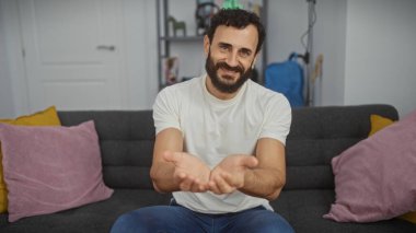 Orta yaşlı, sakallı, gülümseyen, rahat bir oturma odasında ellerini uzatan bir adam.
