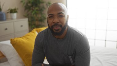 Kapalı bir yatak odasında, içinde sarı yastıklar ve bitkiler olan bir yatakta oturan sakallı yakışıklı genç bir zenci sıcak ve rahat bir ev manzarası yaratıyor..