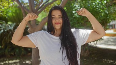 Parktaki bir kadın, kendine güvenerek kollarını açıyor. Arka planda yeşil bir ortamda güç ve vücut pozitifliği sergiliyor. Günlük beyaz bir gömlek giyiyor..