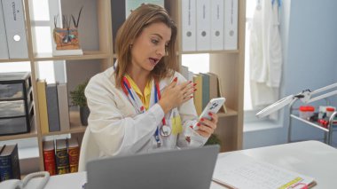 Klinikte çalışan genç, sarışın bir kadın, raflar ve tıbbi dosyalarla çevrili bir yerde steteskoplu beyaz bir laboratuvar önlüğü giyerken akıllı telefondan konuşuyor..