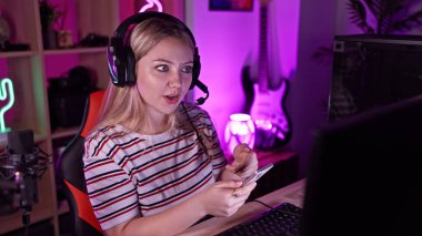 Akşamları neon ışıklı oyun odasında akıllı telefon kullanan kulaklıklı, şaşırmış bir genç kadın..