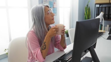 Ofiste kahvesini yudumlayan gri saçlı kadın, görünür dövmeleri olan bir masada oturuyor, modern ve çeşitli iş yerlerini yansıtıyor, bilgisayarlarla ve ofis malzemeleriyle çevrili..