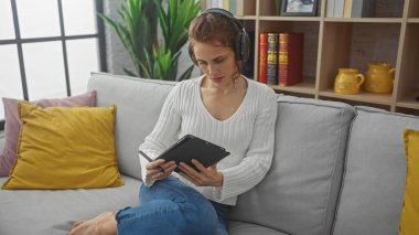 Kulaklık takan genç bir kadın rahat bir oturma odasında kanepede otururken tablet kullanıyor..