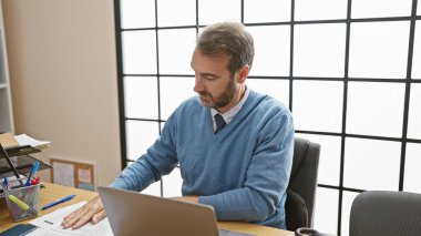Dizüstü bilgisayarı, kağıtları ve pencere ızgarasıyla çalışan orta yaşlı bir adam.