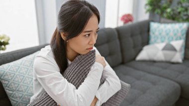 Düşünceli, genç Asyalı bir kadın, dekoratif yastıklarla dolu gri bir kanepede oturur, evde rahat ve düşünceli bir anı canlandırır..