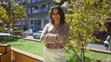 Kollarını kavuşturmuş, genç, güzel bir İspanyol kadın. Güneşli bir şehir parkında duruyor. Arkasında binalar ve arabalar var..