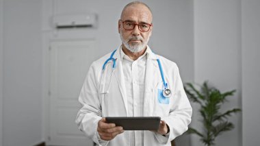 Gri saçlı, sakallı, kendinden emin bir erkek doktor elinde tabletle bir hastane odasında duruyor..