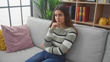 Çizgili süveter giyen genç İspanyol kadın kapalı bir yerdeki gri koltukta düşünceli bir şekilde oturuyor. Etrafı renkli yastıklarla ve bitkilerle çevrili..