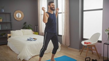 Sakallı bir adam modern bir yatak odasında spor malzemeleriyle kutlar. Yaşam tarzı dengesi ve rahatlığı yansıtır..