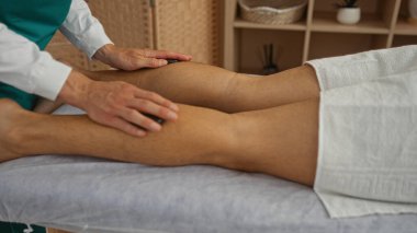 Sağlık merkezinde masaj yaptıran bir adam masör ellerini bacaklarına baskı uygulamak için kullanır ve kapalı alanda rahatlatıcı bir spa ortamı yaratır.