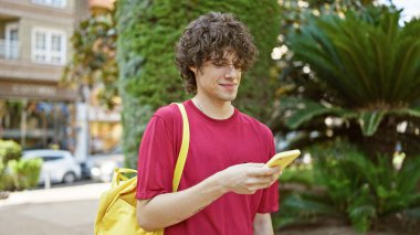 Kıvırcık saçlı, sıradan kıyafetler giyen yakışıklı bir genç. Parkta akıllı telefon kullanırken gülümsüyor..