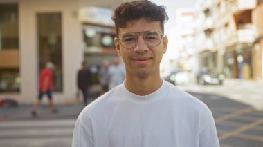 Yakışıklı İspanyol erkek kalabalık bir şehir sokağında duruyor, gözlüklü ve beyaz tişörtlü, binalı ve arka planda insanlar var..