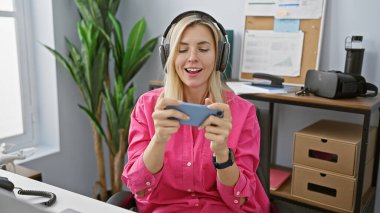 Modern ofiste akıllı telefon kullanan kulaklıklı, gülümseyen bir kadın.