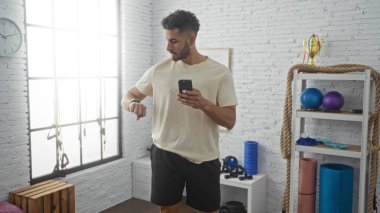 Yakışıklı İspanyol genç adam, arka planında ekipman ve kupalarla modern bir spor salonunda elinde telefonla akıllı saatini kontrol ediyor..