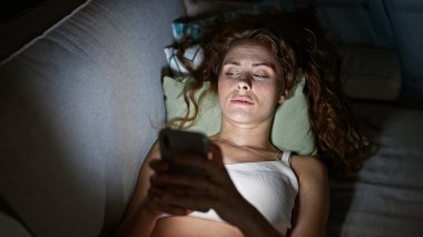 Kafkasyalı genç bir kadın gece vakti akıllı telefonunu tutarak yatağında uzanıyor ve modern bir yaşam tarzı yaratıyor..