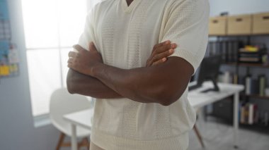 Arka planda modern beyaz bir masa olan bir ofis odasında kolları çapraz duran Afrikalı Amerikalı genç bir adam.