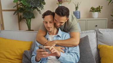 Birbirini seven iki adam evdeki bir koltukta otururken, biri cep telefonu kullanırken diğeri ona arkadan sarılıyor, rahat yastıklar ve oturma odasındaki bitkilerle çevrili..