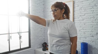 Spor salonunda dambıllarıyla egzersiz yapan yetişkin bir İspanyol kadın, spor merkezinde spor yaparken spor programını sergiliyor..