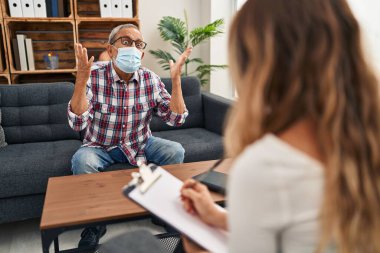 Gri saçlı neşeli son sınıf öğrencisi, tıbbi maske takarak kapalı bir klinikte oturuyor, neşeli bir gülümseme ve olumlu bir ifadeyle zaferi kutluyor.