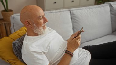 Beyaz sakallı, evde akıllı telefon kullanırken rahat bir oturma odasındaki kanepede dinlenen yaşlı kel adam..