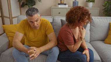 Orta yaşlı bir adam ve bir kadın oturma odasında kanepede oturuyor, üzgün ve mesafeli görünüyorlar, bir çiftin evdeki ciddi ilişki sorununu resmediyorlar..
