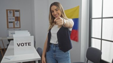 Genç, çekici, sarışın bir kadın, arka planda Venezuela bayrağı olan kapalı bir üniversite odasında dikilirken doğrudan kamerayı işaret ediyor..