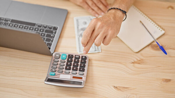 Close-up of a man's hand using a calculator next to a laptop and us dollars on a wooden table in an office setting.