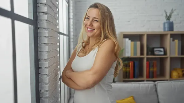 stock image Young caucasian woman with crossed arms, smiling in a bright cozy living room with a bookshelf background.